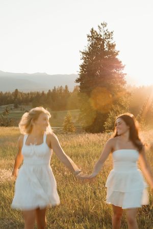 Two people in white dresses run through a sunlit meadow at golden hour with mountains and trees in the background.