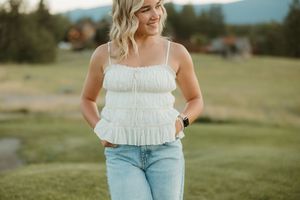 A person in a white ruched tank top and light jeans stands in a grassy field with mountains in the background.