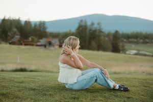 Person in white top and jeans sits thoughtfully on grassy hillside with mountains and rural landscape in background.
