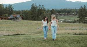 Two people walk together on a grassy hill with mountains and forest in the background on a cloudy day.