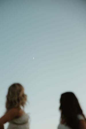 Silhouettes of two people against pale blue sky with tiny crescent moon visible.
