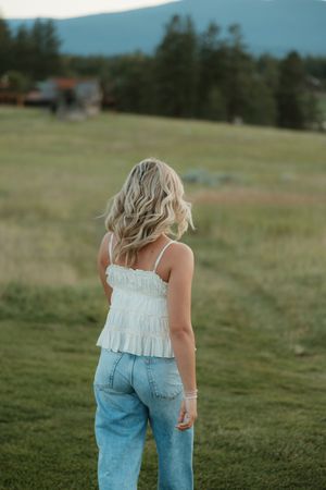 Person wearing white ruffle tank top and blue jeans stands in grassy field with mountains and forest in background.