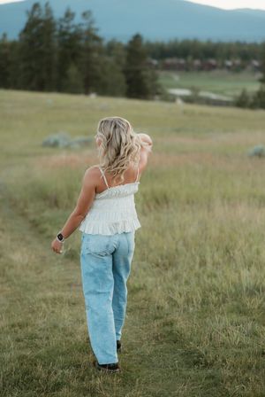 Figure walking through a grassy meadow wearing a white top and light denim jeans with mountains in the background.