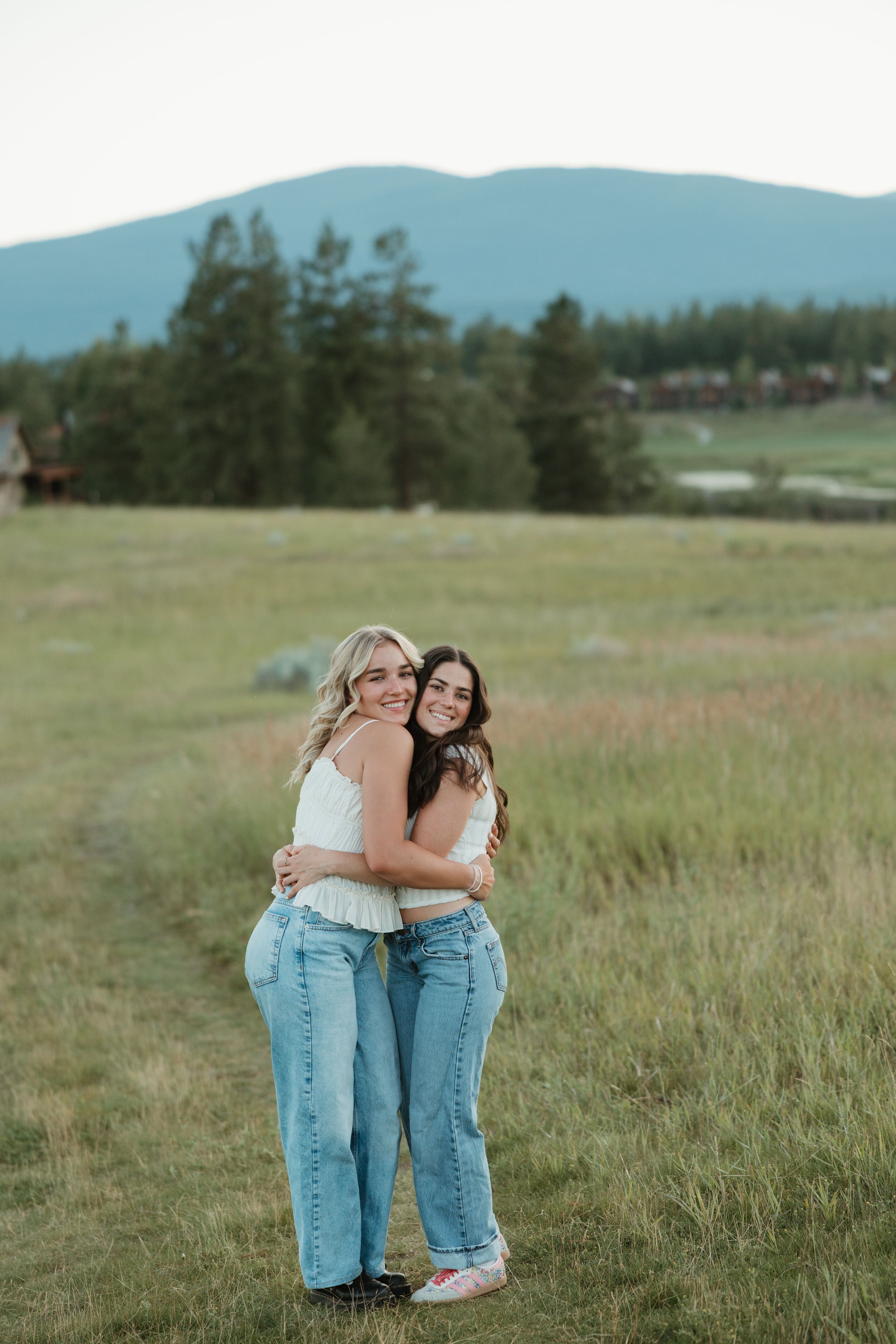 Two friends share a warm embrace while standing in a grassy field with mountains and pine trees in the background.