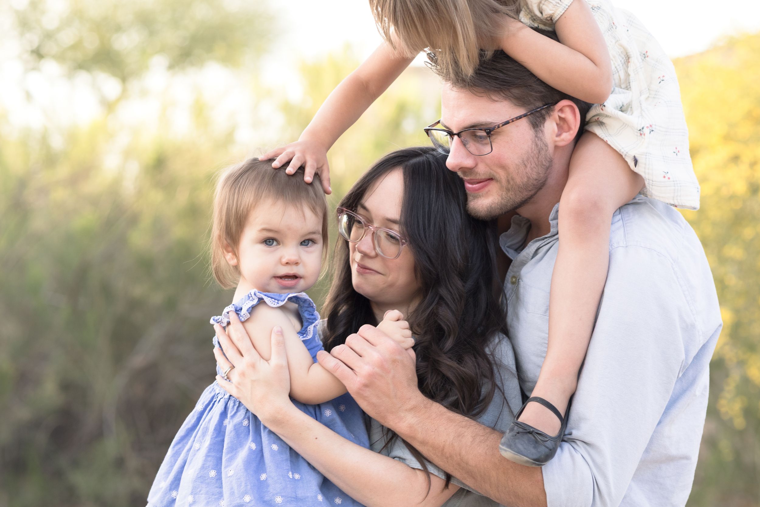 Family of four with mom holding baby and dad cuddled in behind her while toddler is on his shoulders in front of flowers in Queen Creek,