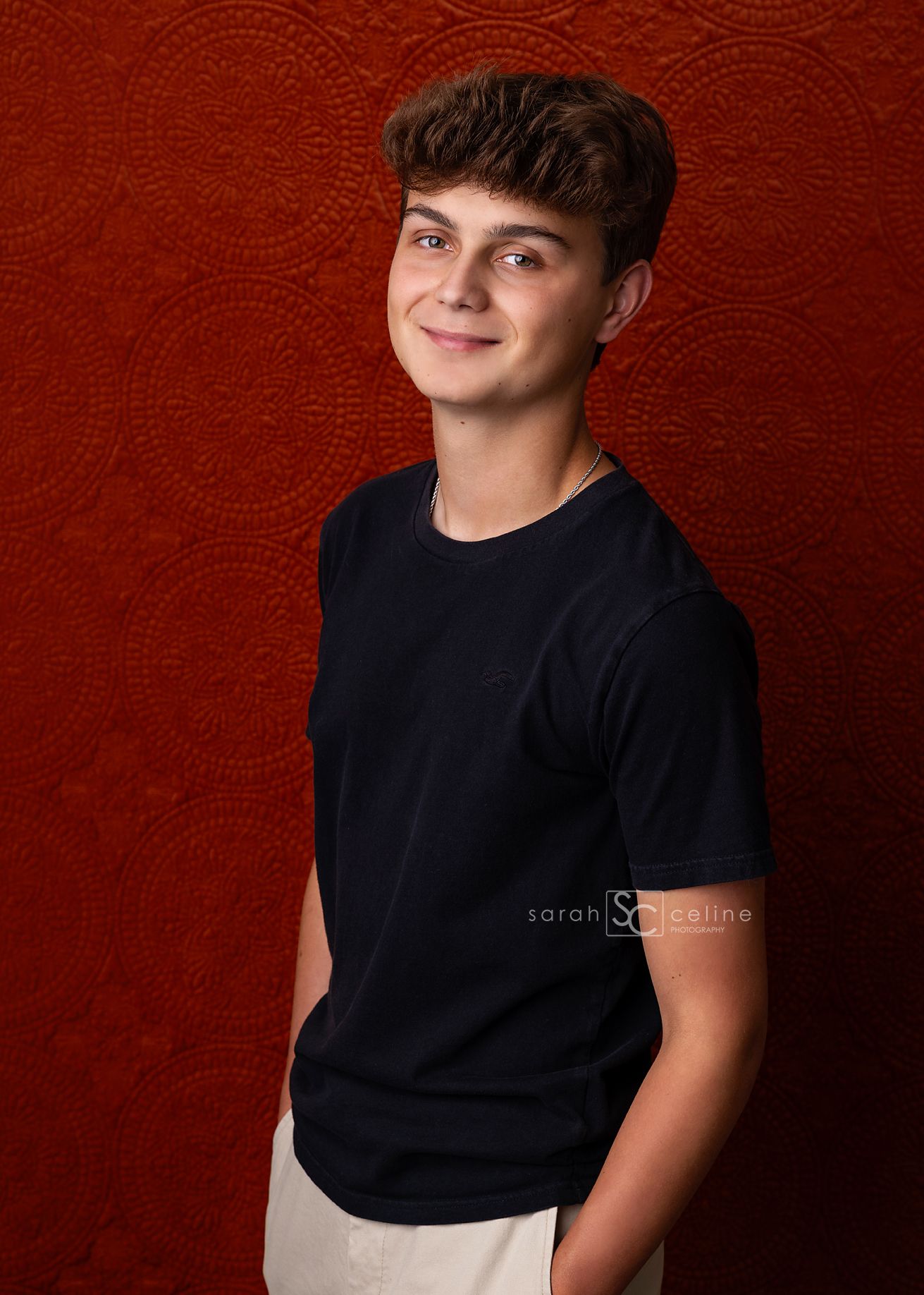 Young person in a black t-shirt poses against a deep red background in a casual studio portrait series.