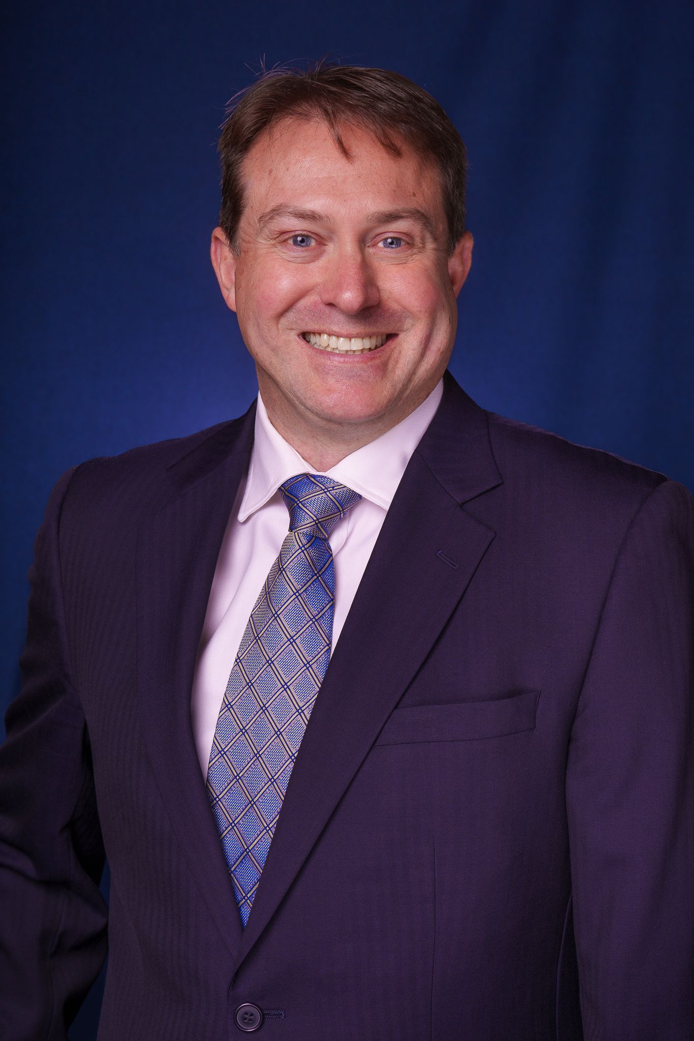 Professional male executive in navy suit and blue tie smiling for corporate headshot on dark blue background.