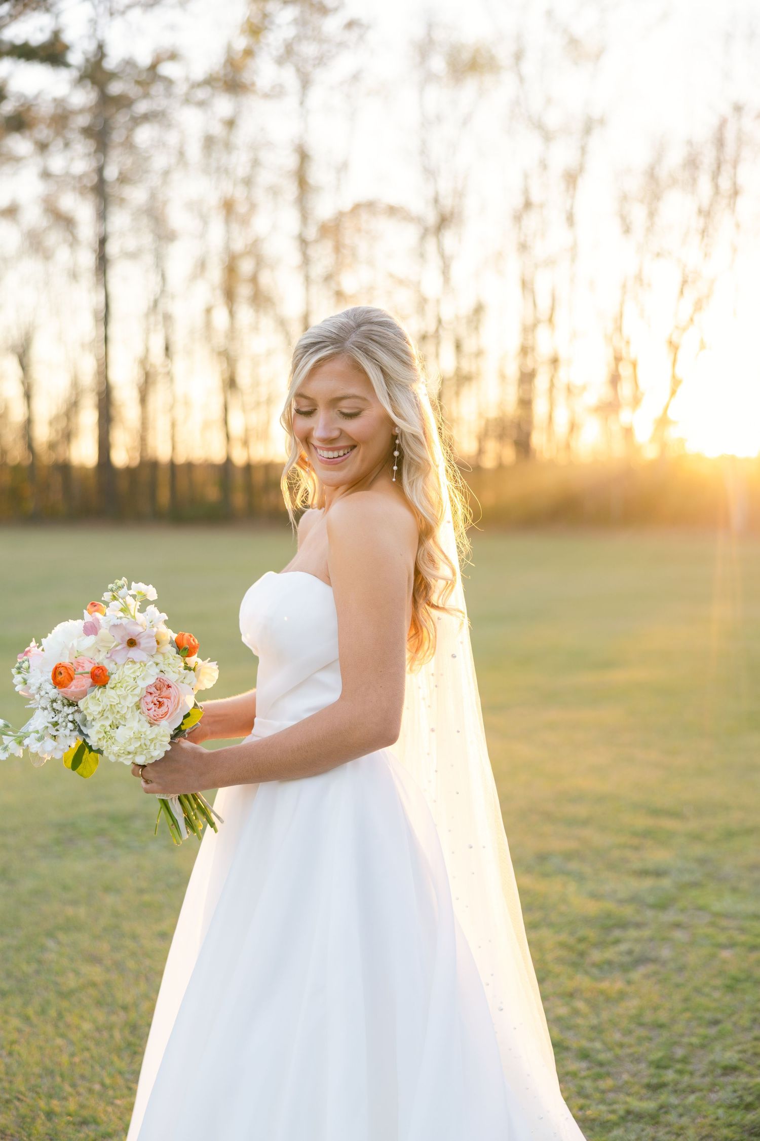 Bride in flowing white wedding dress holds bouquet of white and coral flowers at golden sunset in open field.