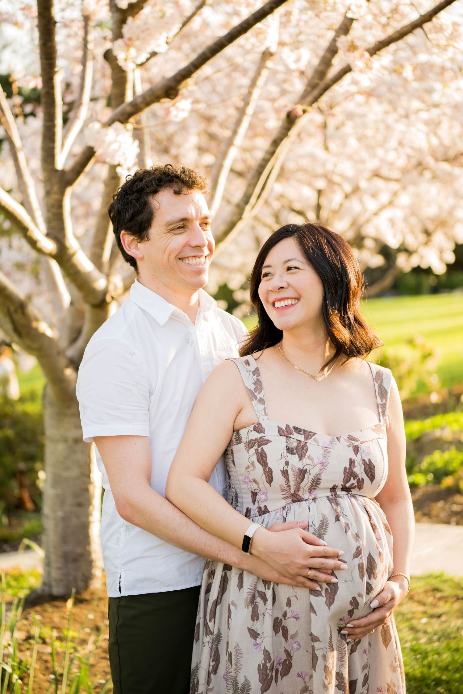 Pregnant woman in a floral maxi dress smiling at her partner, hands on her belly, under blossoming cherry trees at Piedmont Park.