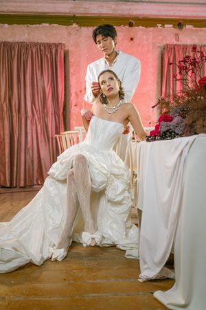A couple in a romantic wedding photoshoot pose against pink walls and curtains in an intimate indoor setting.