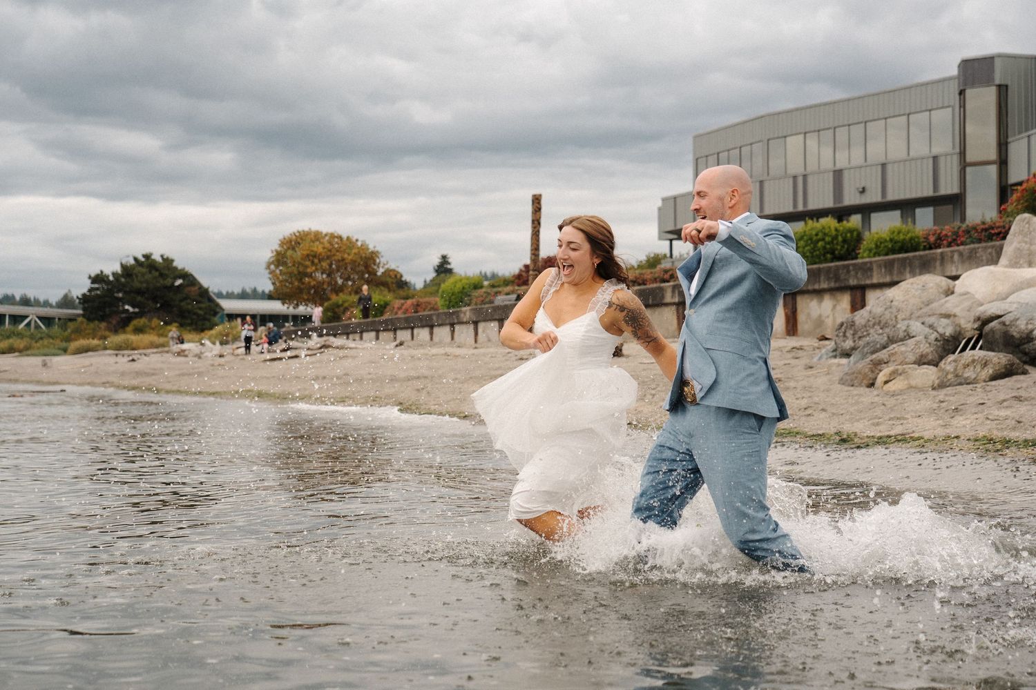 Newlywed couple splashes through shallow water at the beach in their wedding attire during a playful photoshoot.