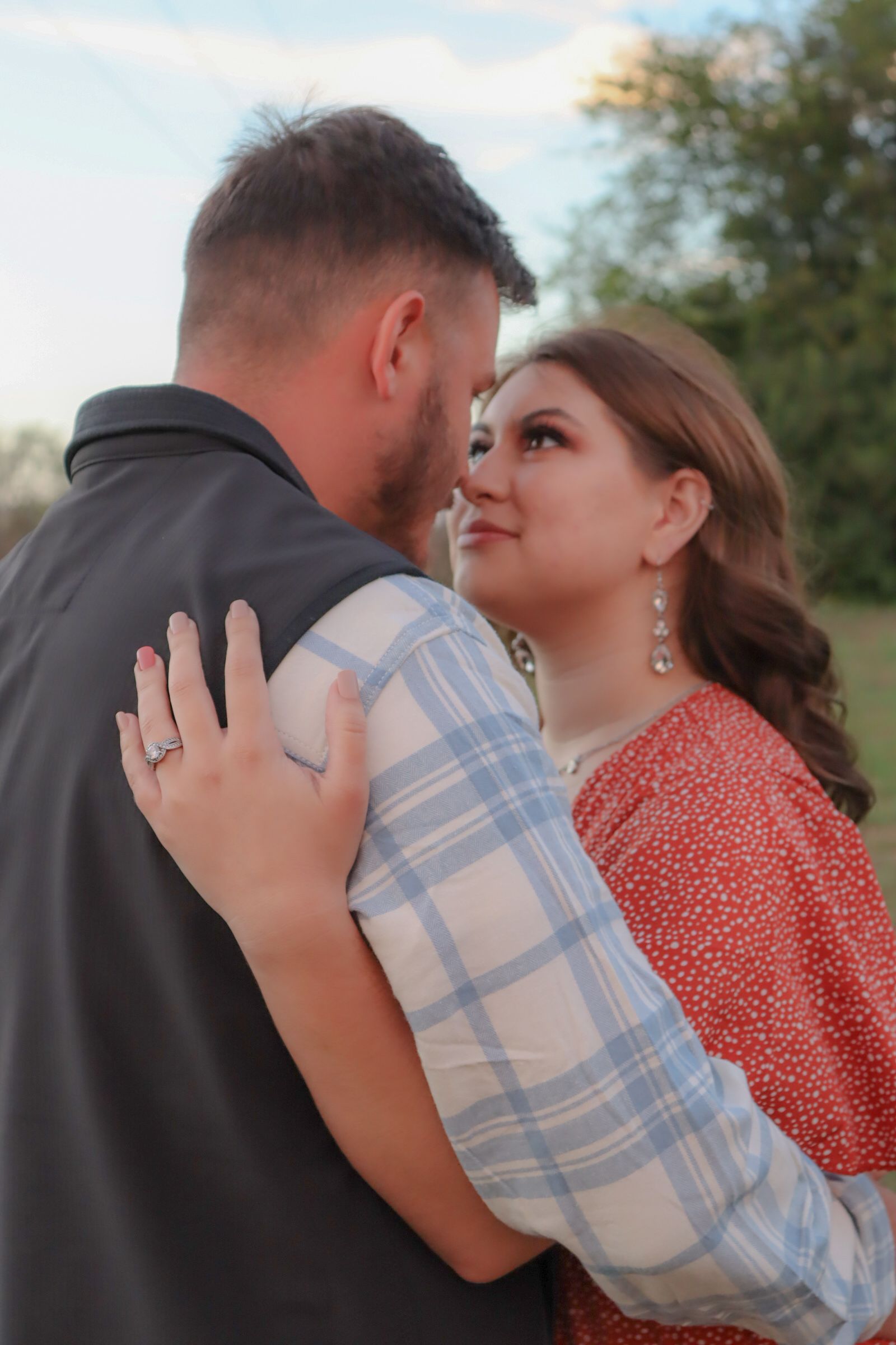 A romantic outdoor engagement photo features a couple embracing, with the woman wearing a red dress and sparkling earrings.