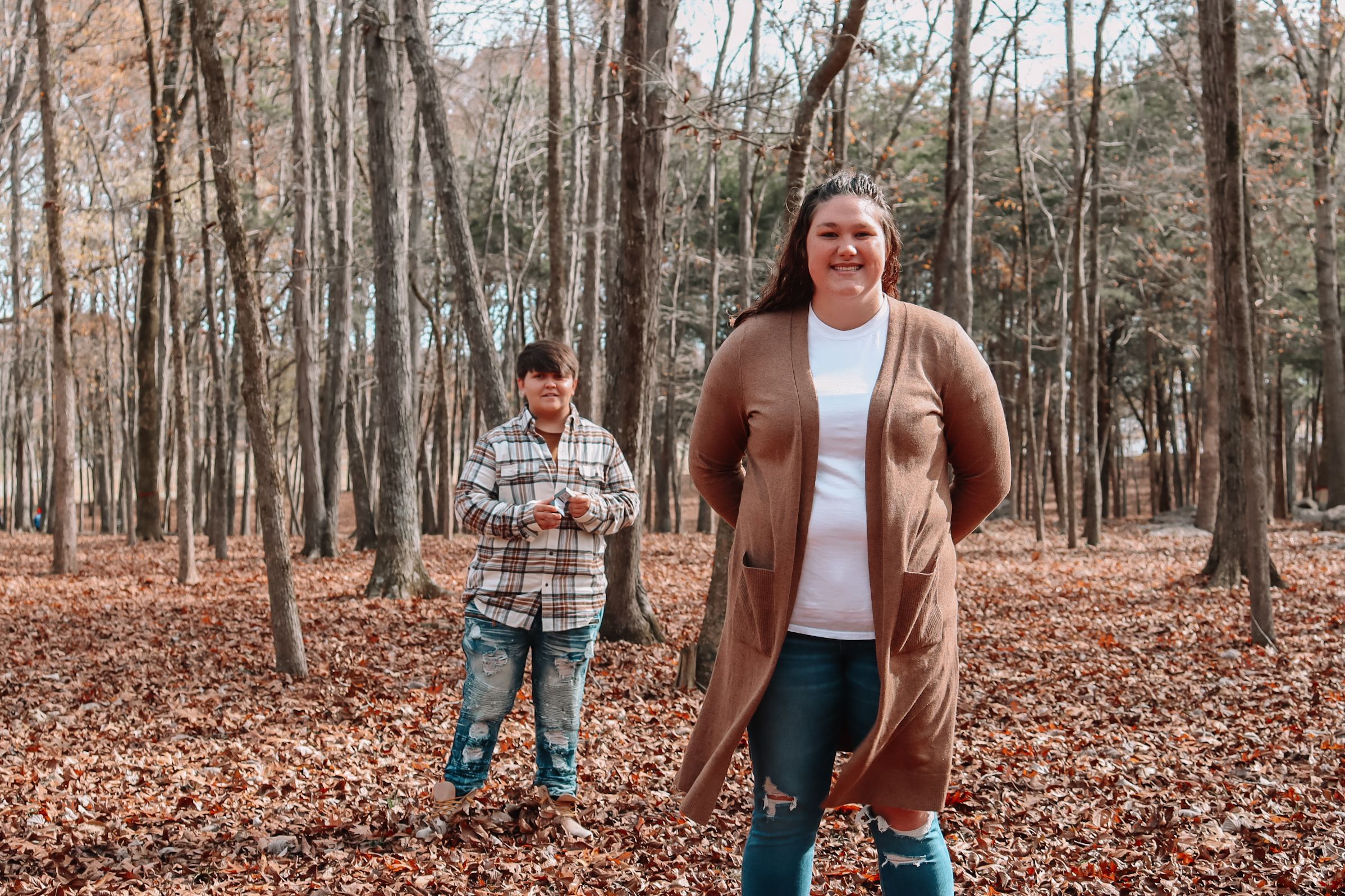Two people stand in a wooded forest with fallen autumn leaves covering the ground while wearing casual winter attire.