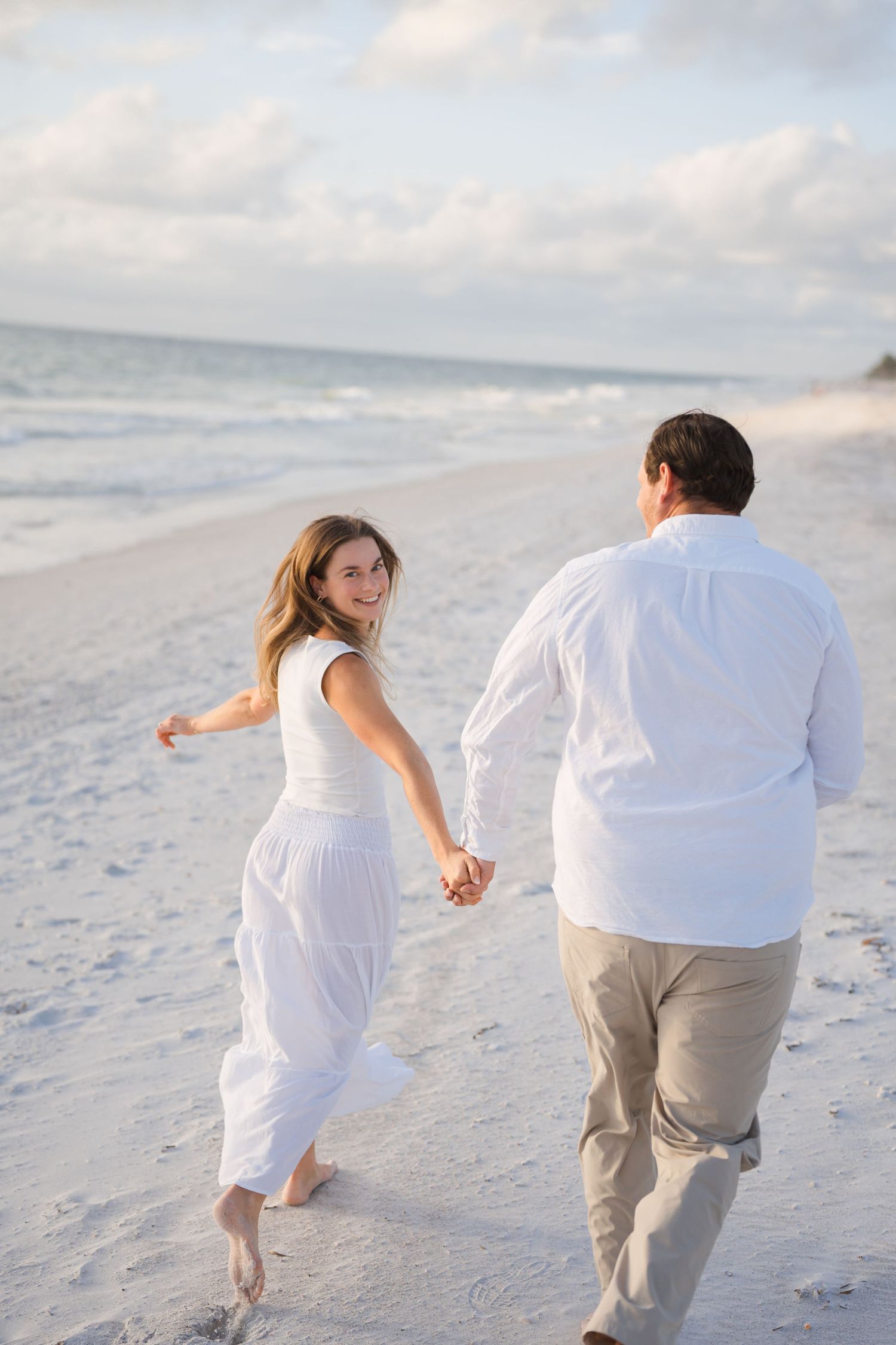 Couple in white attire walks hand in hand along pristine beach at sunset.