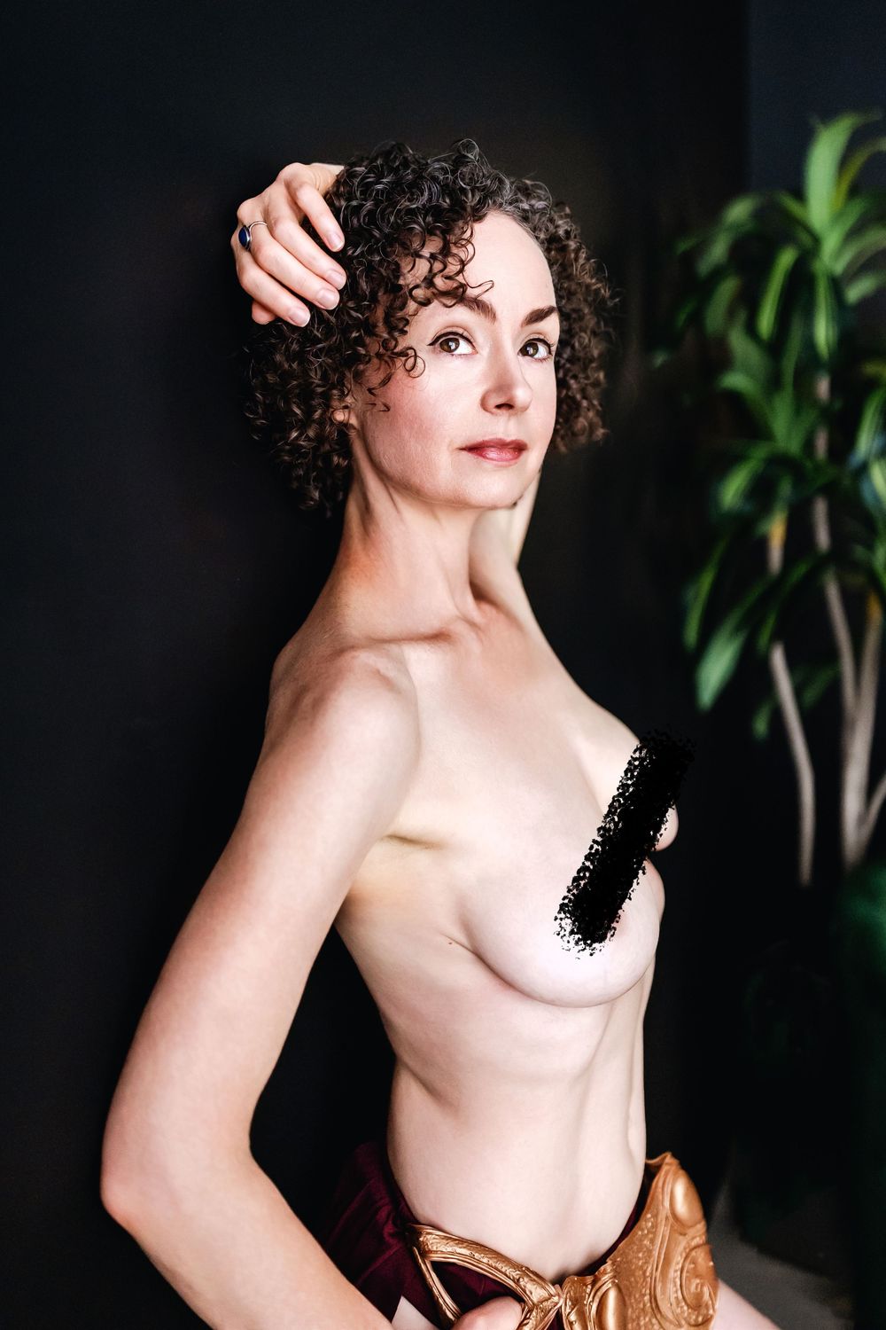 A person with curly dark hair poses in an artistic portrait with a partial black top against a dark background.