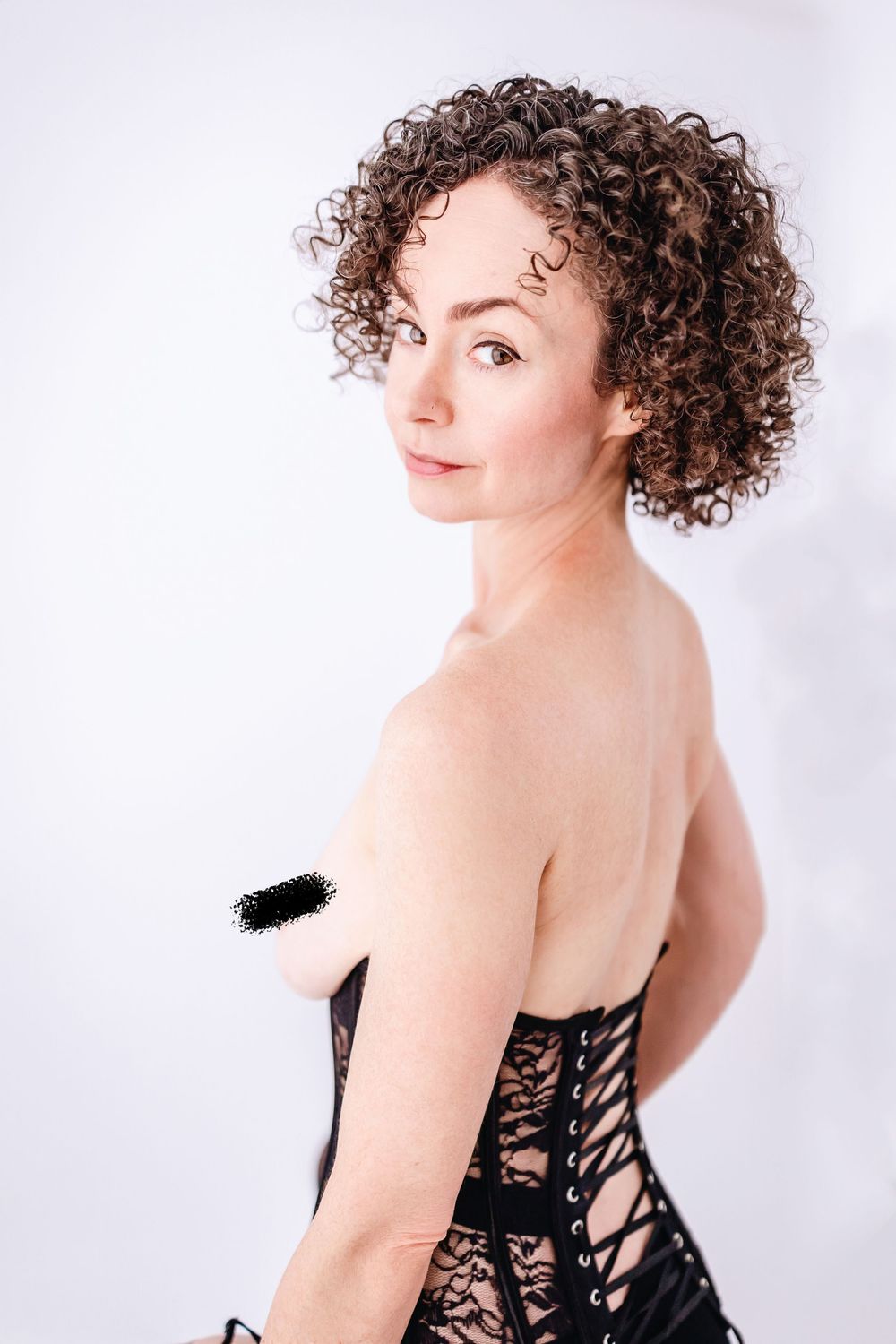 Portrait of a model with natural curly hair wearing black lace corset against white background.