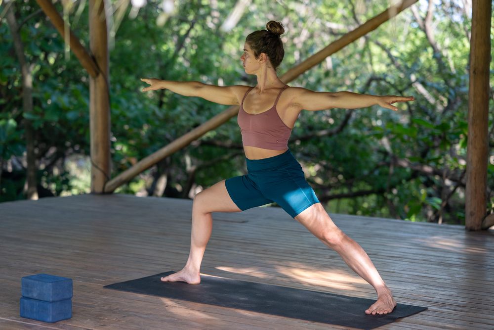 Someone practicing yoga in warrior pose on an outdoor deck with a yoga block and mat in a lush garden setting.