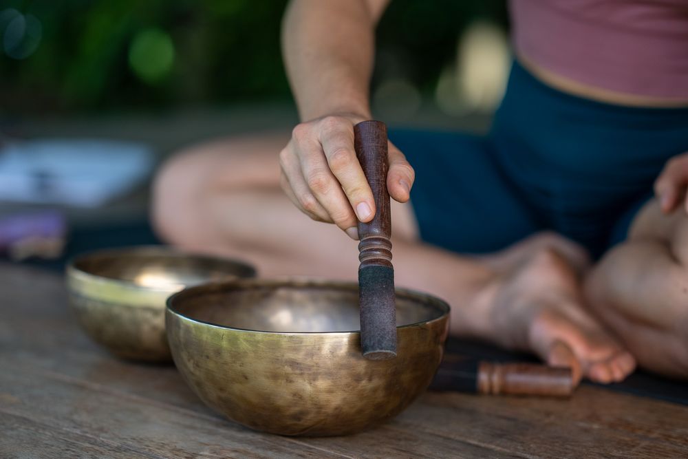 Multiple hands strike singing bowls with mallets in sequence, creating a meditative soundscape in a serene setting.