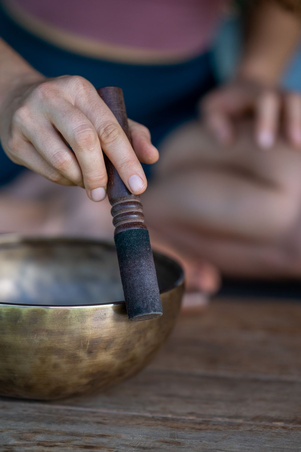 A hand holds a wooden mallet while playing a Tibetan singing bowl on a rustic wooden surface.