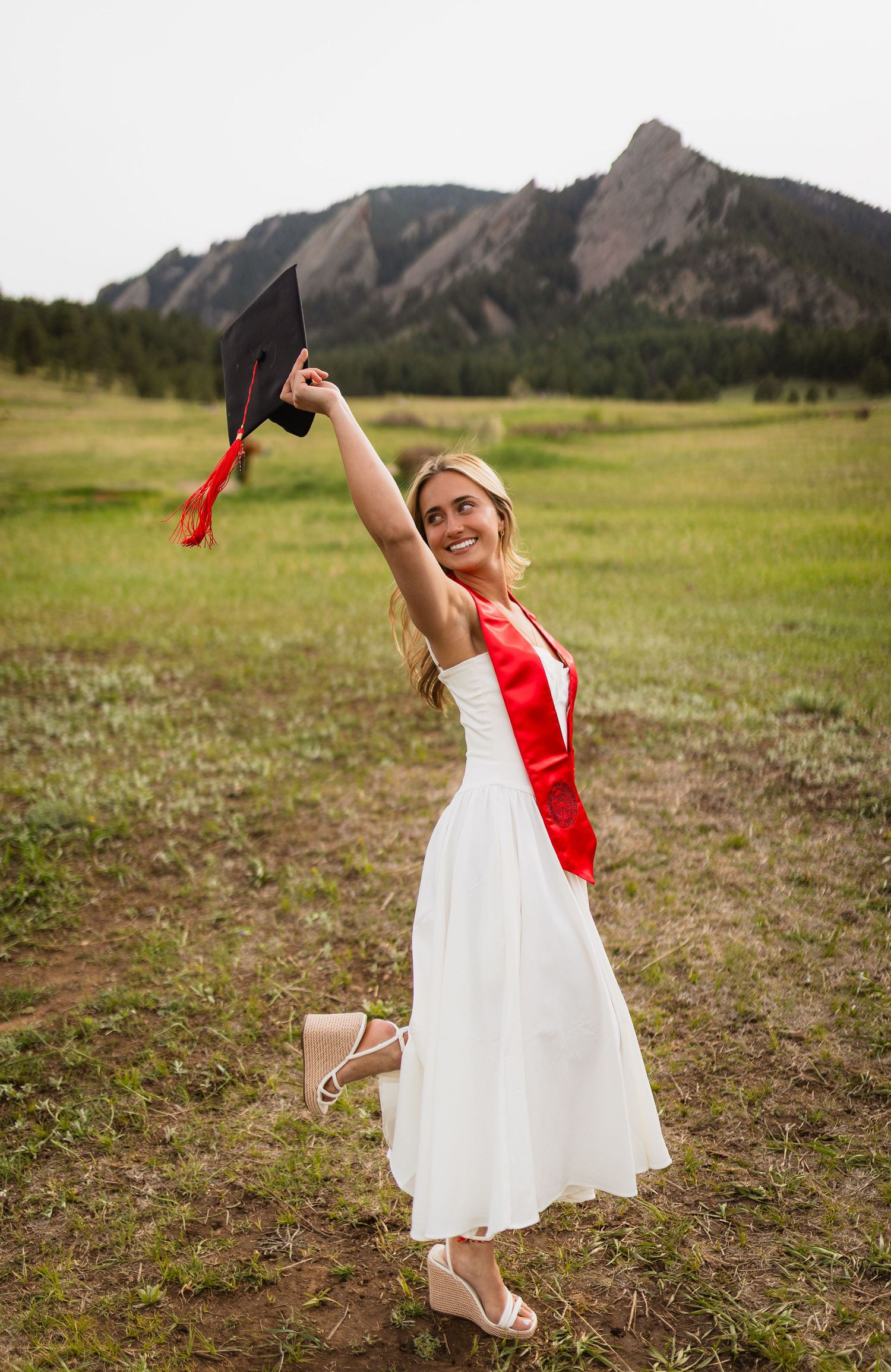 A graduate in a white dress and red sash celebrates with their cap against a backdrop of rocky mountains in Boulder, Colorado.
