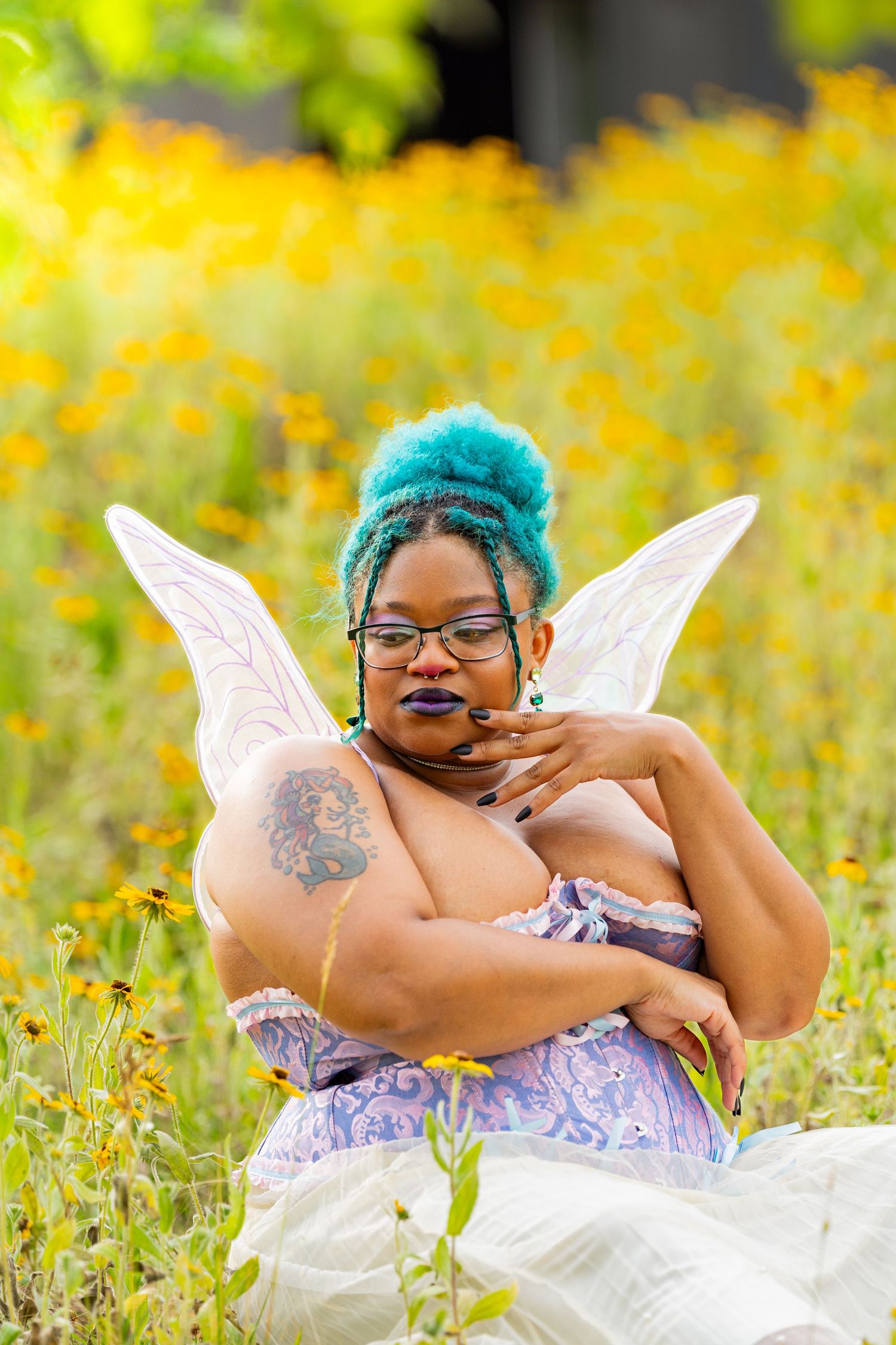 A person with teal hair wearing fairy wings poses playfully in a field of yellow wildflowers.