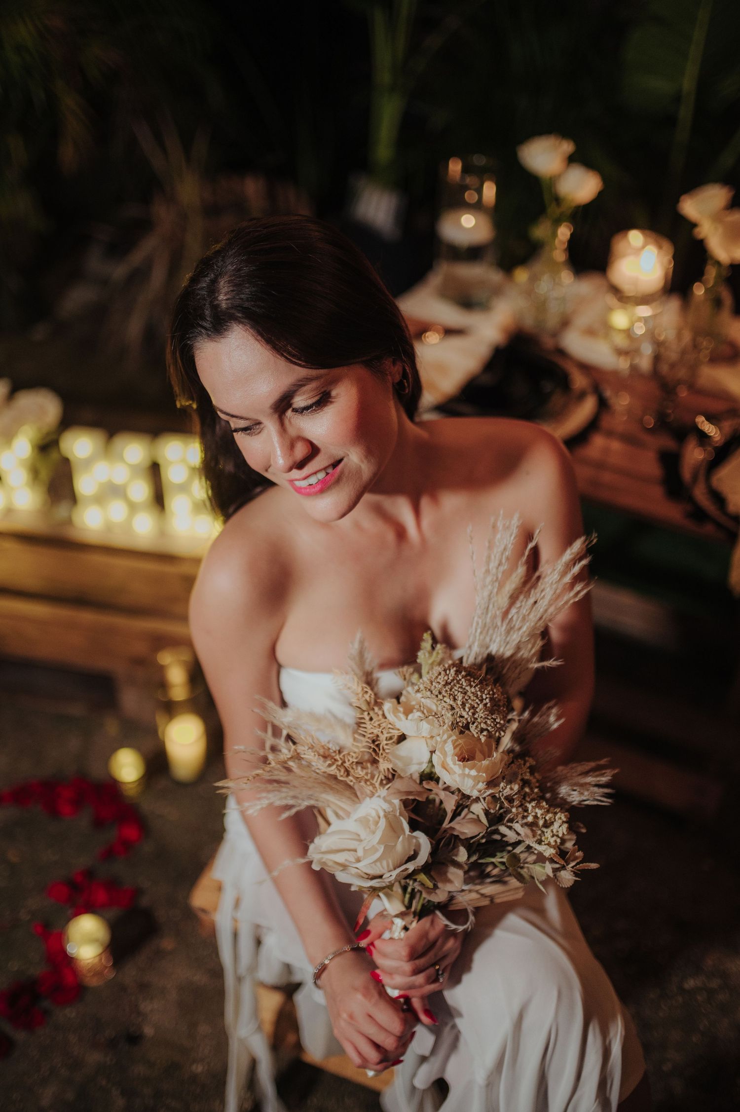 A bride in a white dress holds a romantic bouquet of dried flowers and roses in warm evening lighting.
