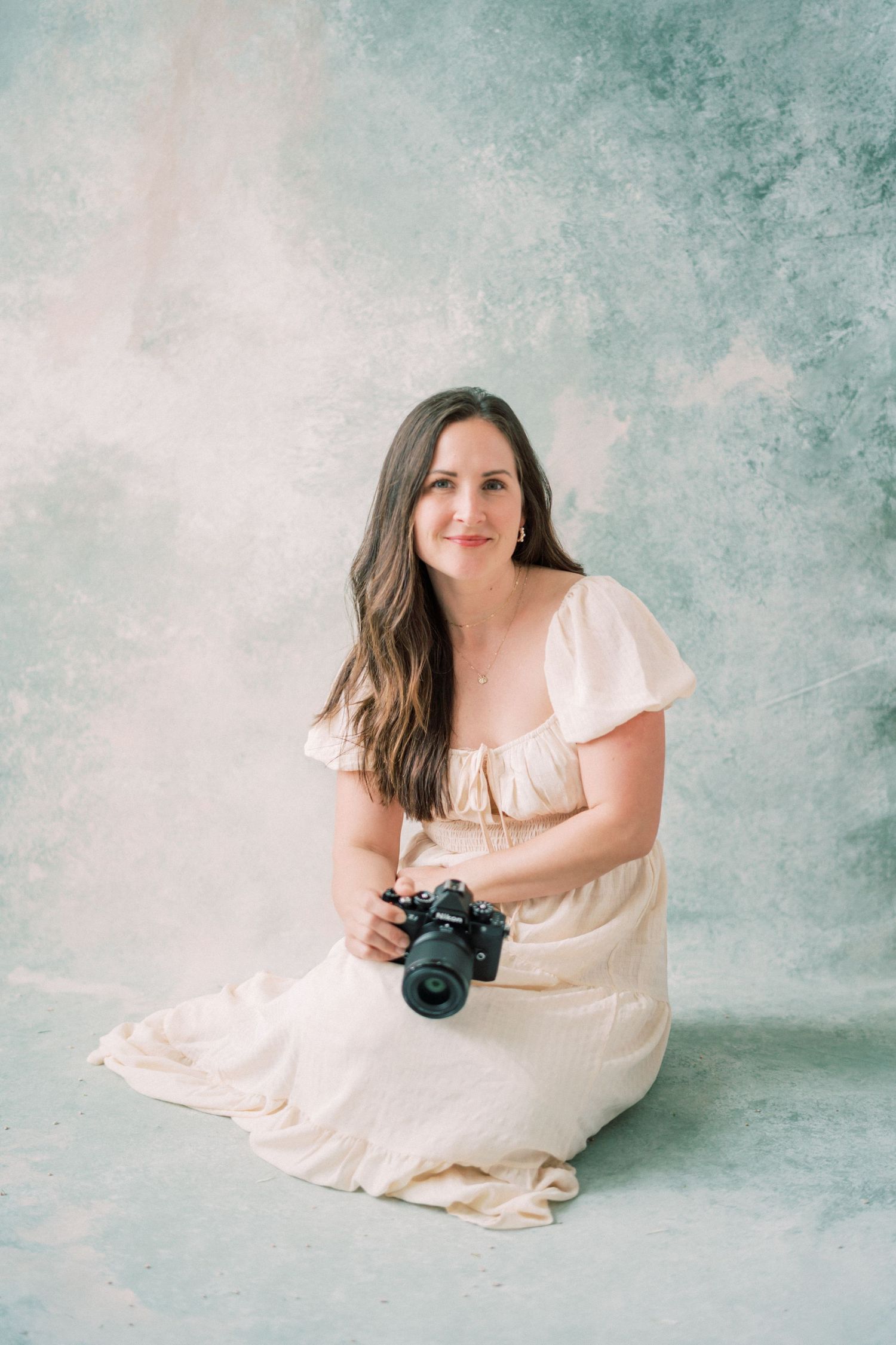 Professional photographer in black blazer and jeans sitting on beige couch holding camera in bright interior setting.