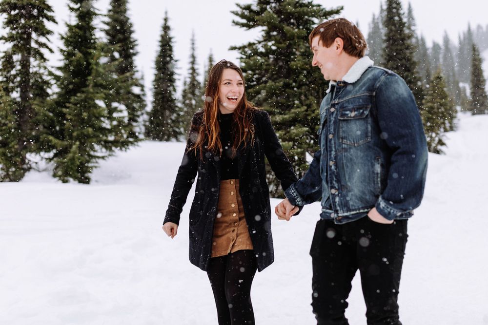 A couple smiling and walking through a snowy forest landscape with evergreen trees in winter.