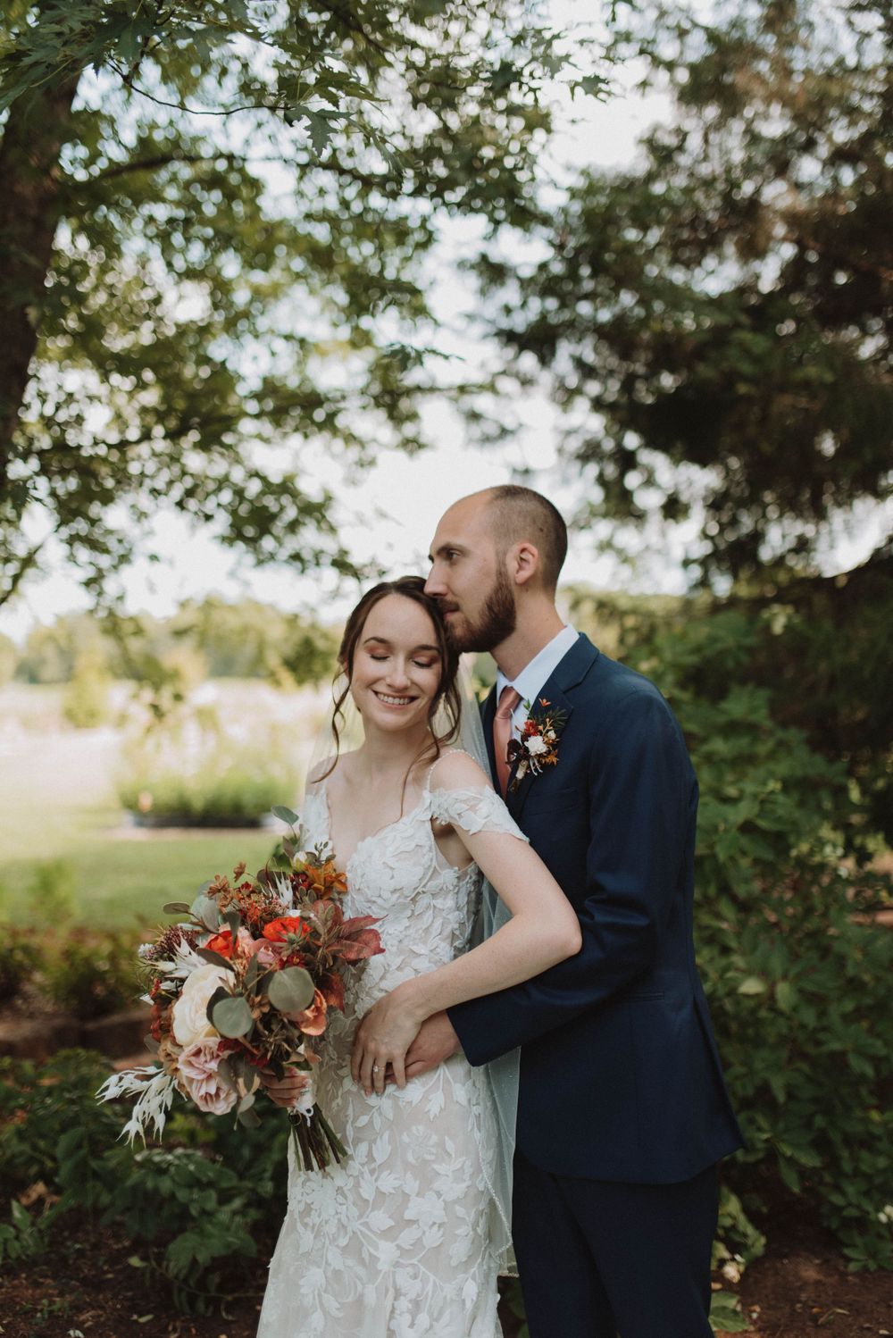 A couple in wedding attire embrace outdoors among trees while holding a vibrant autumn-colored bouquet.