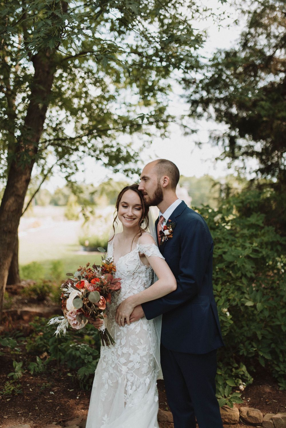 A bride in a lace gown holds an orange and burgundy fall-themed bouquet while posing with her groom in a navy suit outdoors.