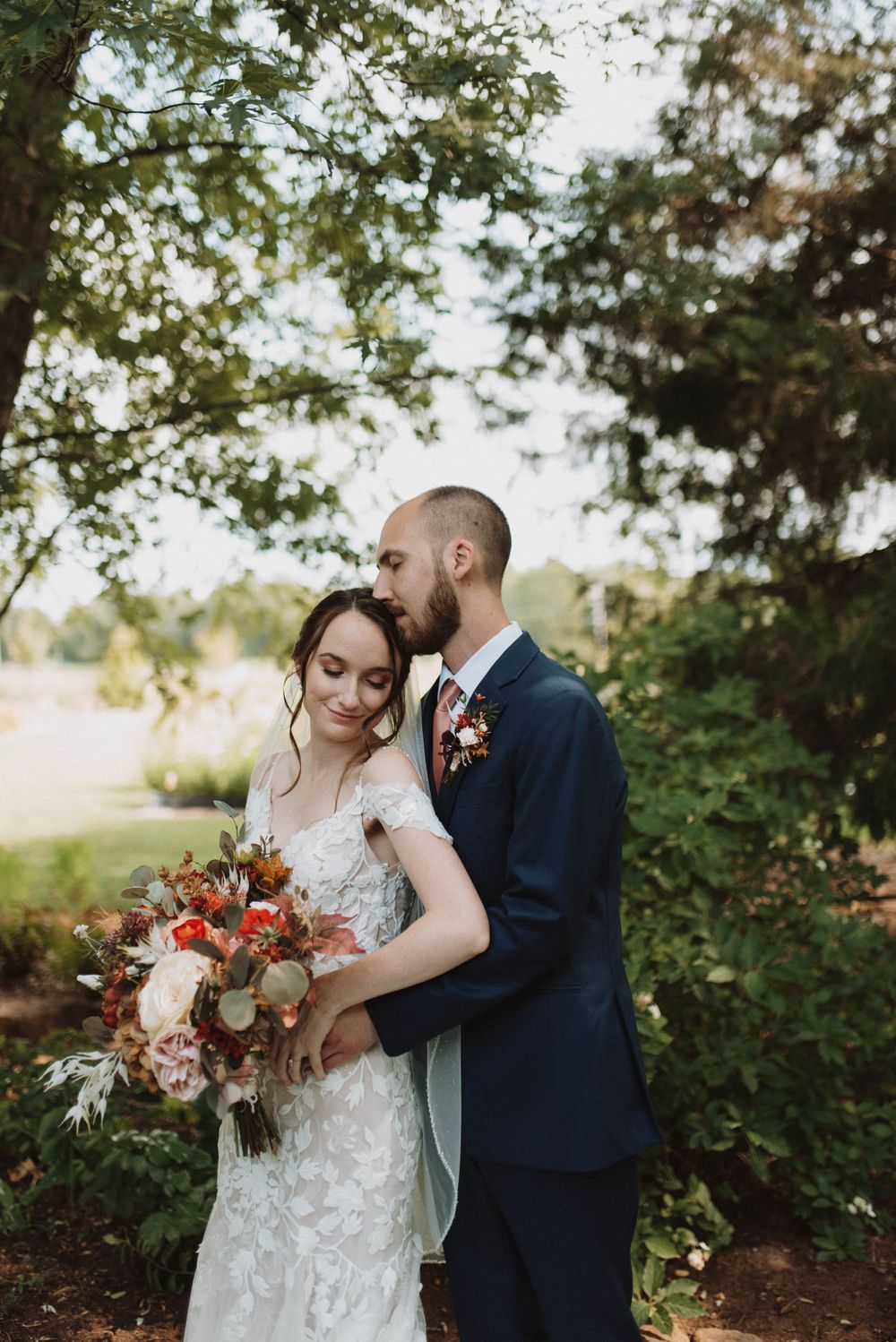 Romantic outdoor wedding portrait with autumn themed bouquet featuring orange and white florals in a natural setting.