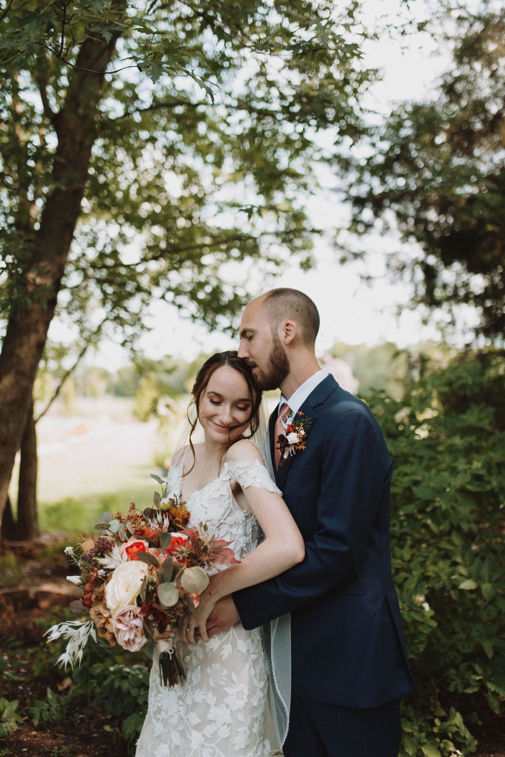 A romantic wedding photo of a couple in a wooded setting with the bride holding a vibrant autumn-colored bouquet.
