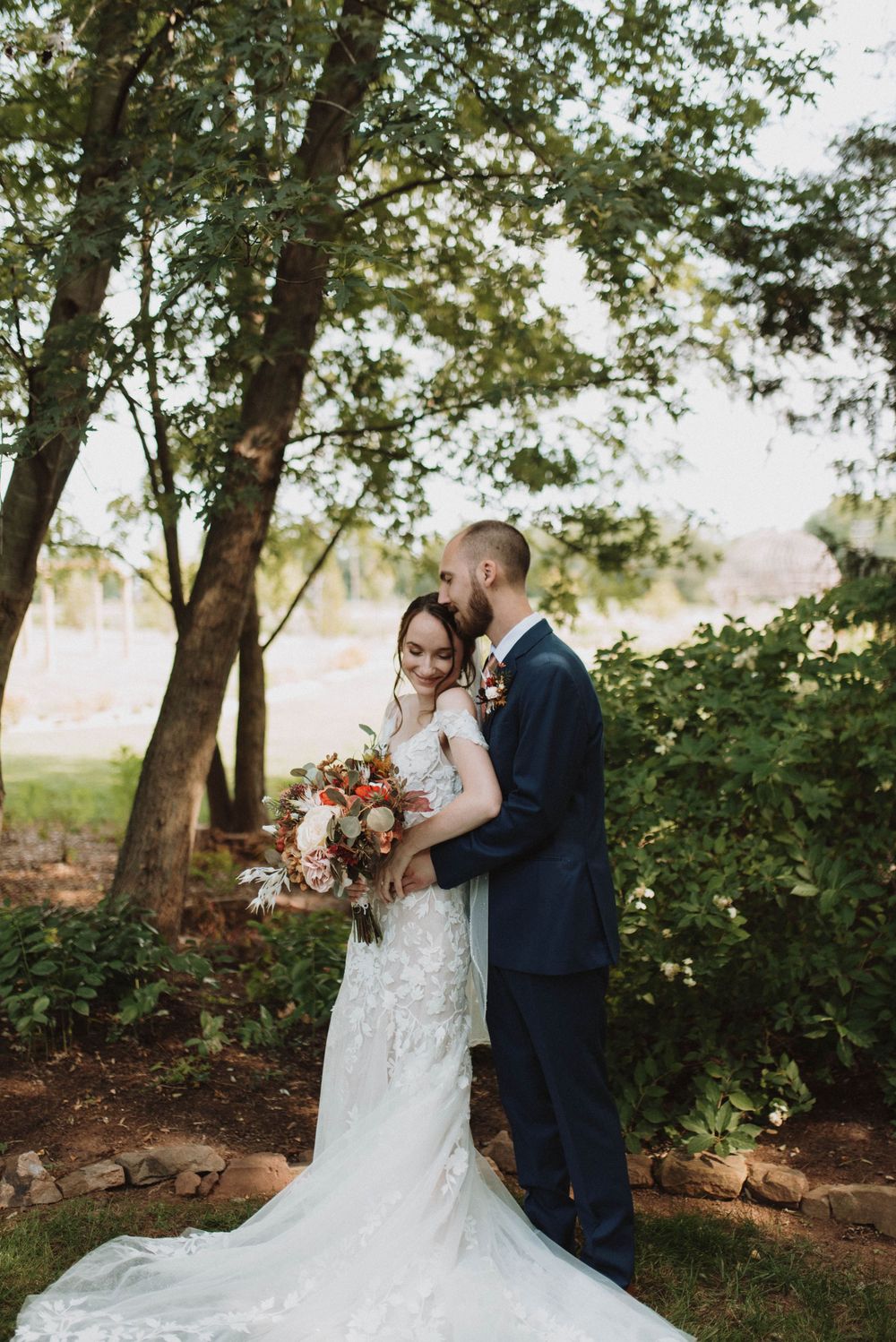 A newlywed couple shares an intimate moment among trees while the bride's lace dress train flows behind her.