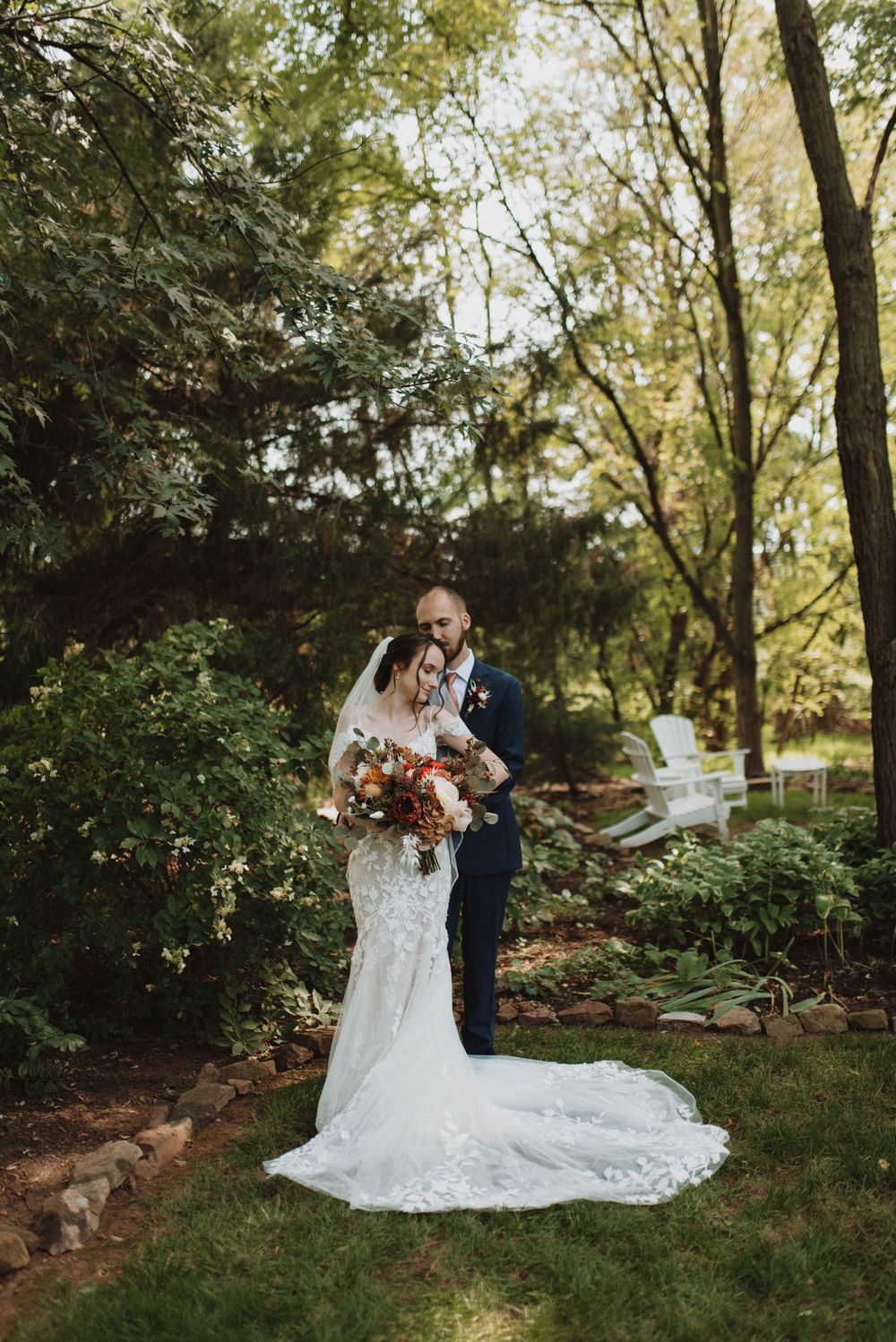 A wedding couple embraces in a romantic garden setting surrounded by lush greenery and natural sunlight.