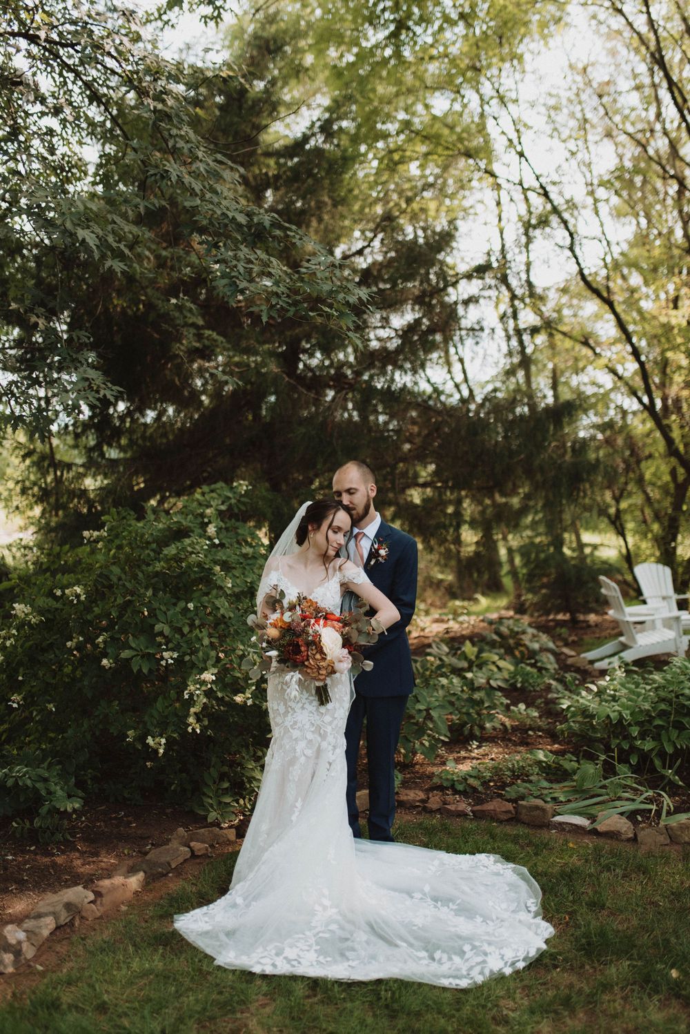 A newlywed couple embraces in a romantic garden setting with fall foliage and a stunning lace wedding gown with long train.