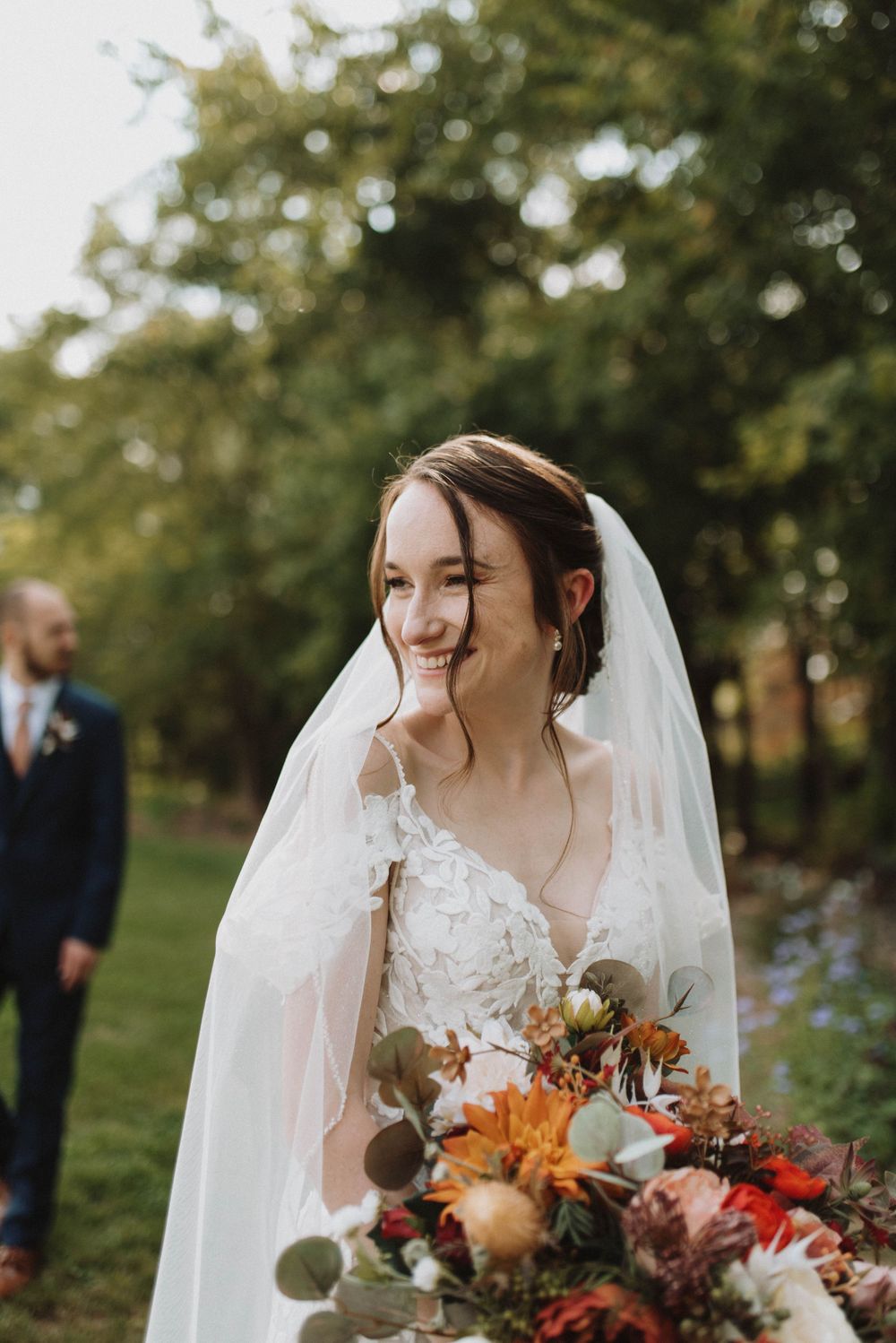 A bride in a lace wedding dress holds an autumn bouquet with orange and burgundy flowers in an outdoor garden setting.