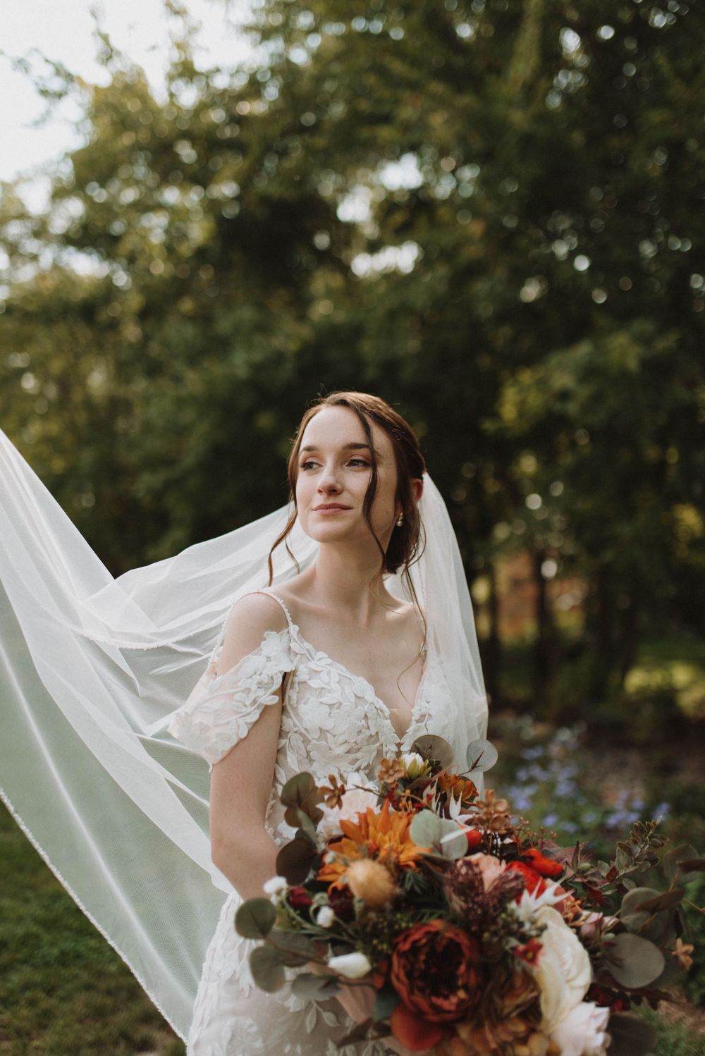 Bride in delicate off-shoulder wedding gown holds vibrant autumn-colored bouquet against blurred garden background.
