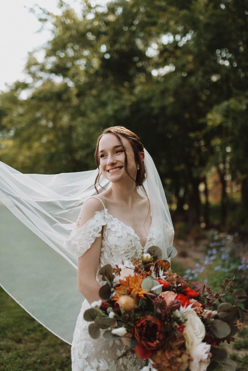 A bride in a lace wedding dress smiles while holding an autumnal bouquet with orange and red flowers.