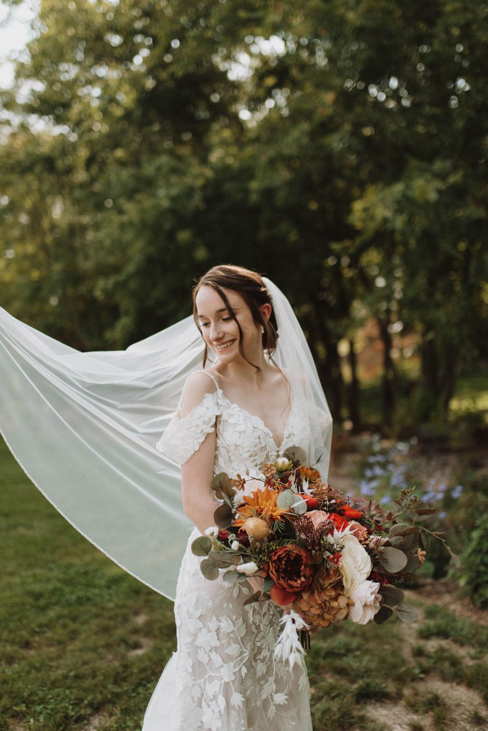 A bride in a lace dress holds a vibrant fall-colored bouquet with her veil flowing in the breeze against a natural backdrop.