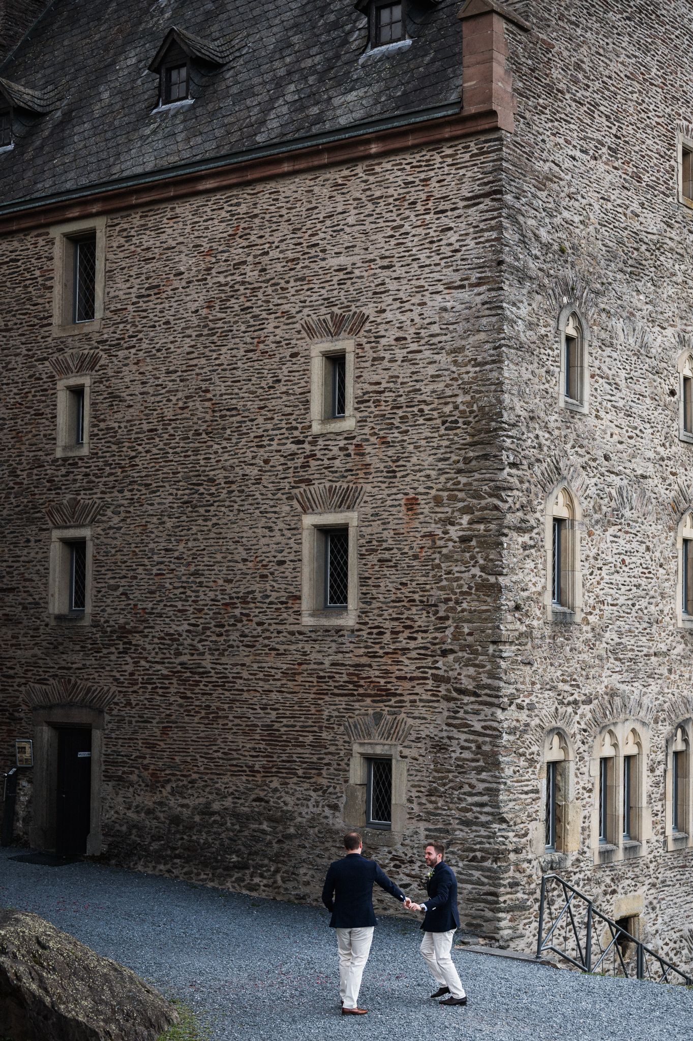 Two people walk along a gravel path next to a historic stone castle wall with small windows in medieval architecture style.