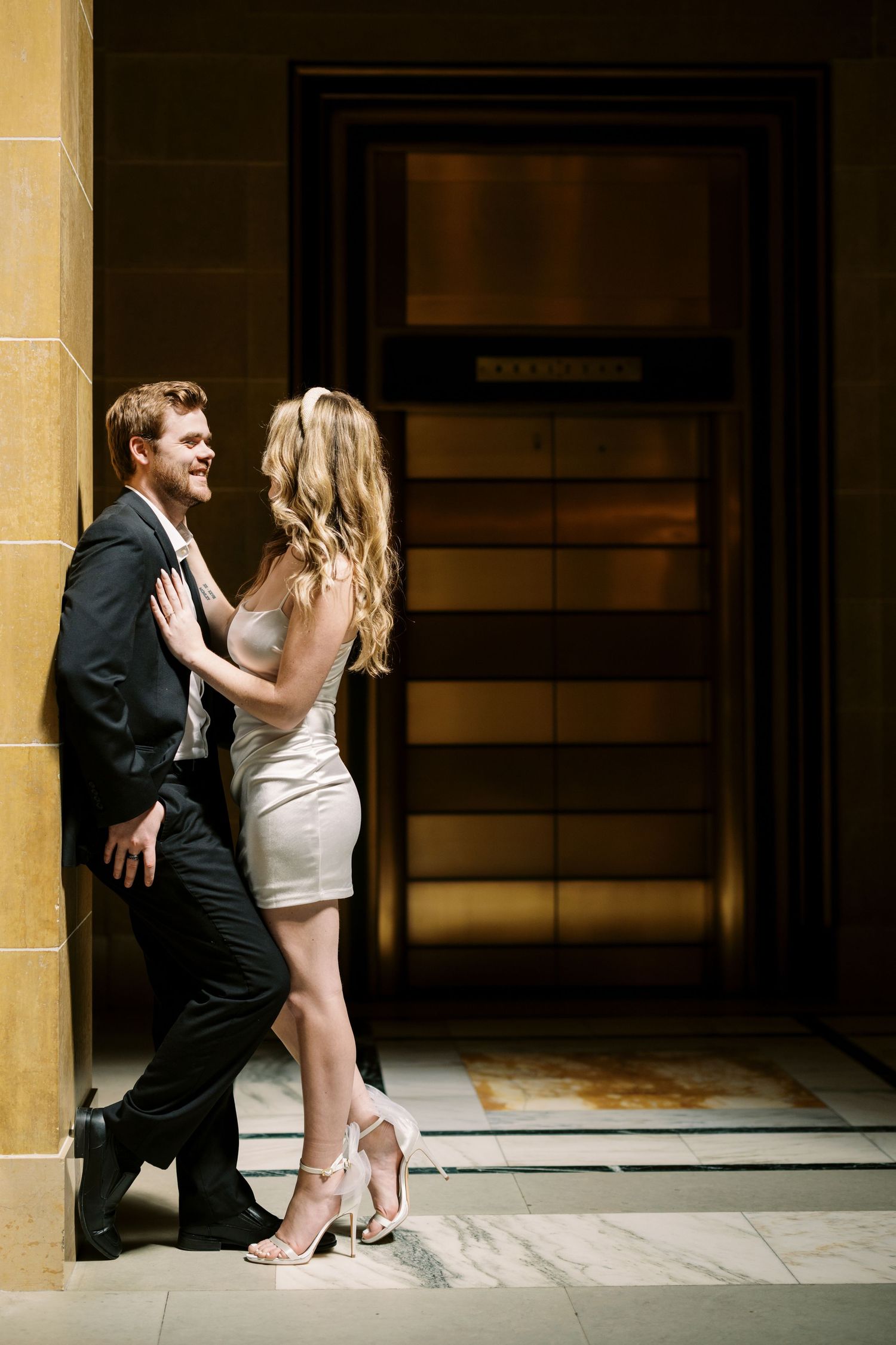A couple in formal attire share an intimate moment by an elevator door.