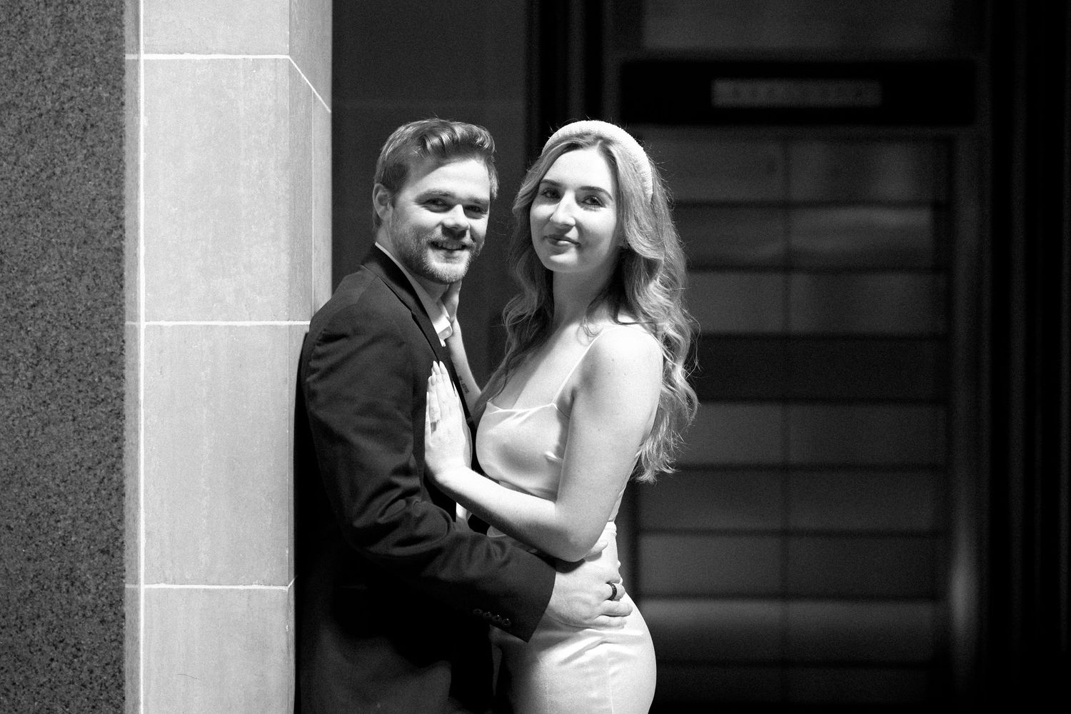 Black and white wedding photo of couple embracing intimately against stone wall in formal attire.