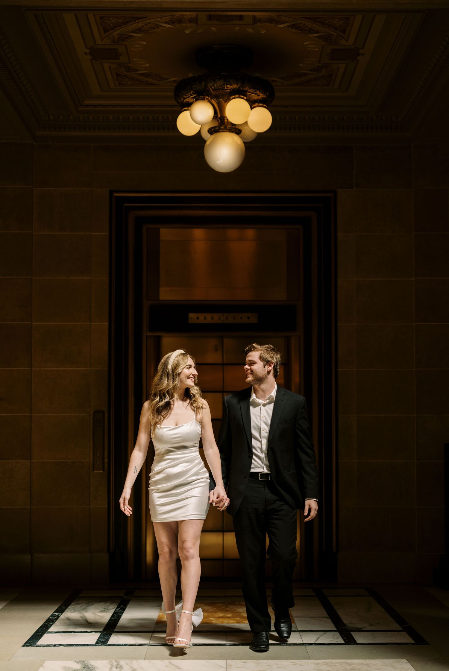 Couple walking through elegant hallway with ornate ceiling light and stone walls in formal evening attire.