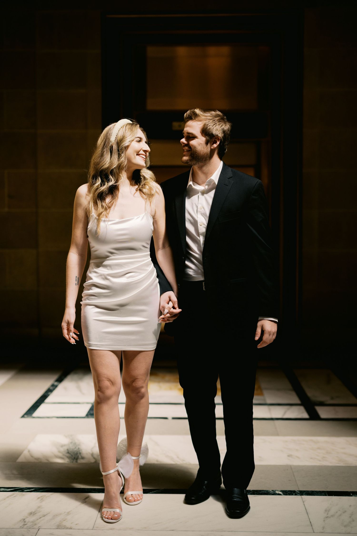 Elegant couple in evening attire holds hands while walking through a dimly lit hallway.