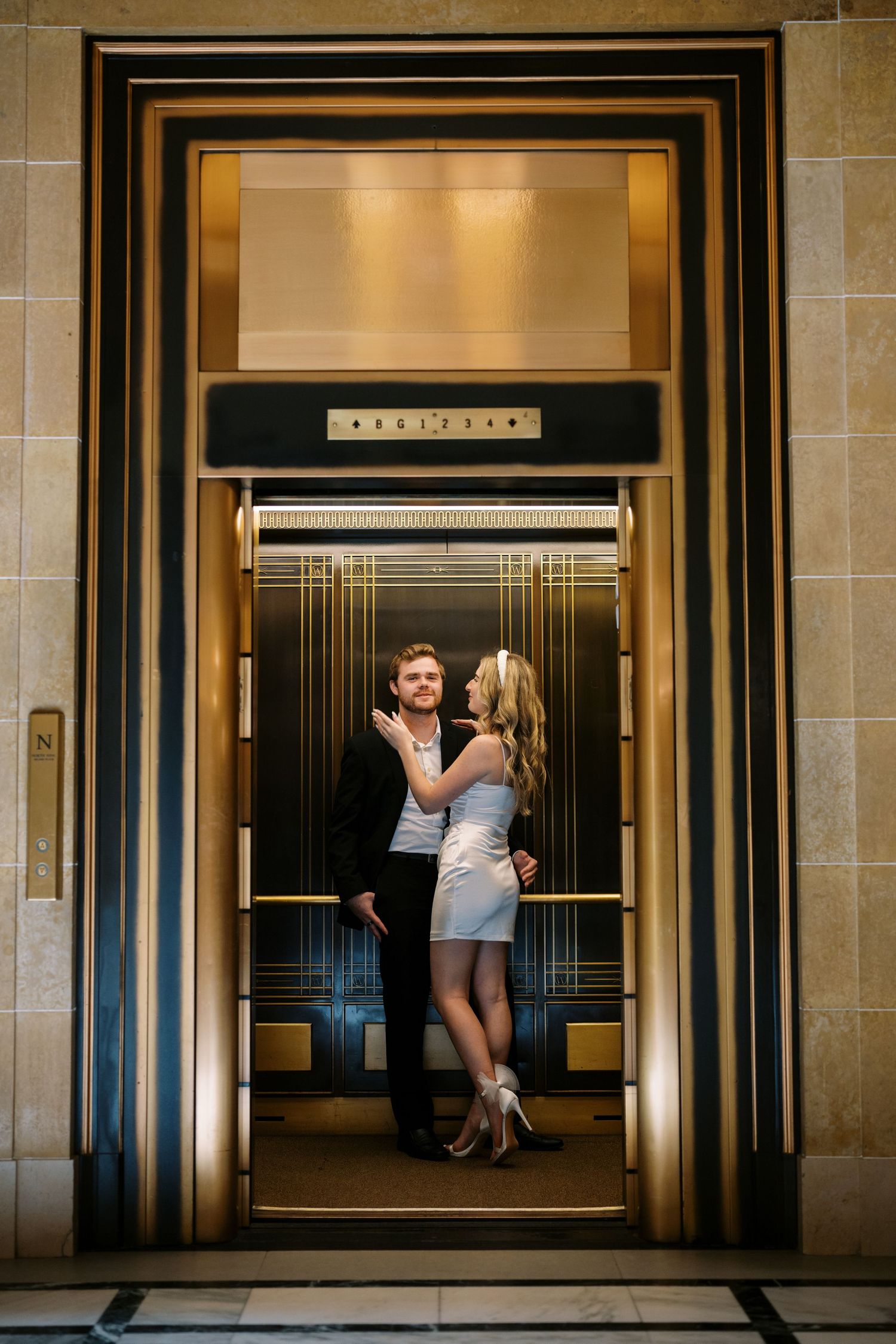 A couple in formal attire shares a romantic moment inside an art deco elevator with gold trim detailing.