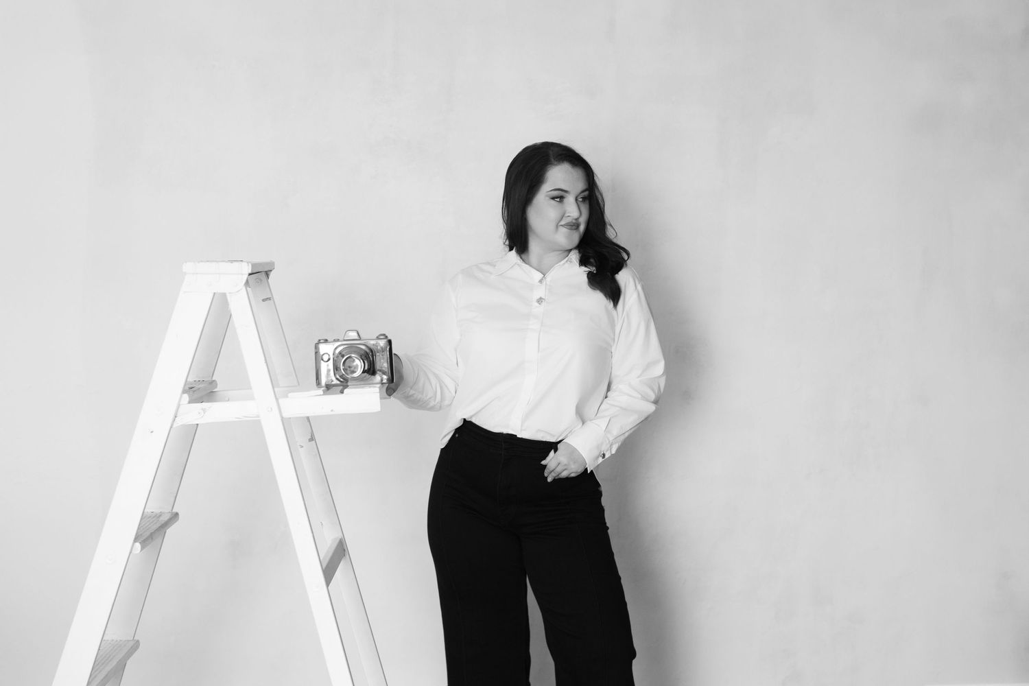 Black and white studio portrait with person standing next to white ladder against plain background.