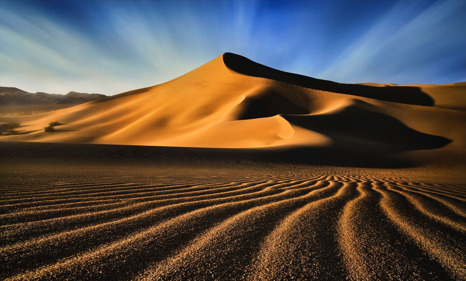Golden sand dunes cast dramatic shadows against a blue sky in a desert landscape during sunset by Evan Will in Death Valley California