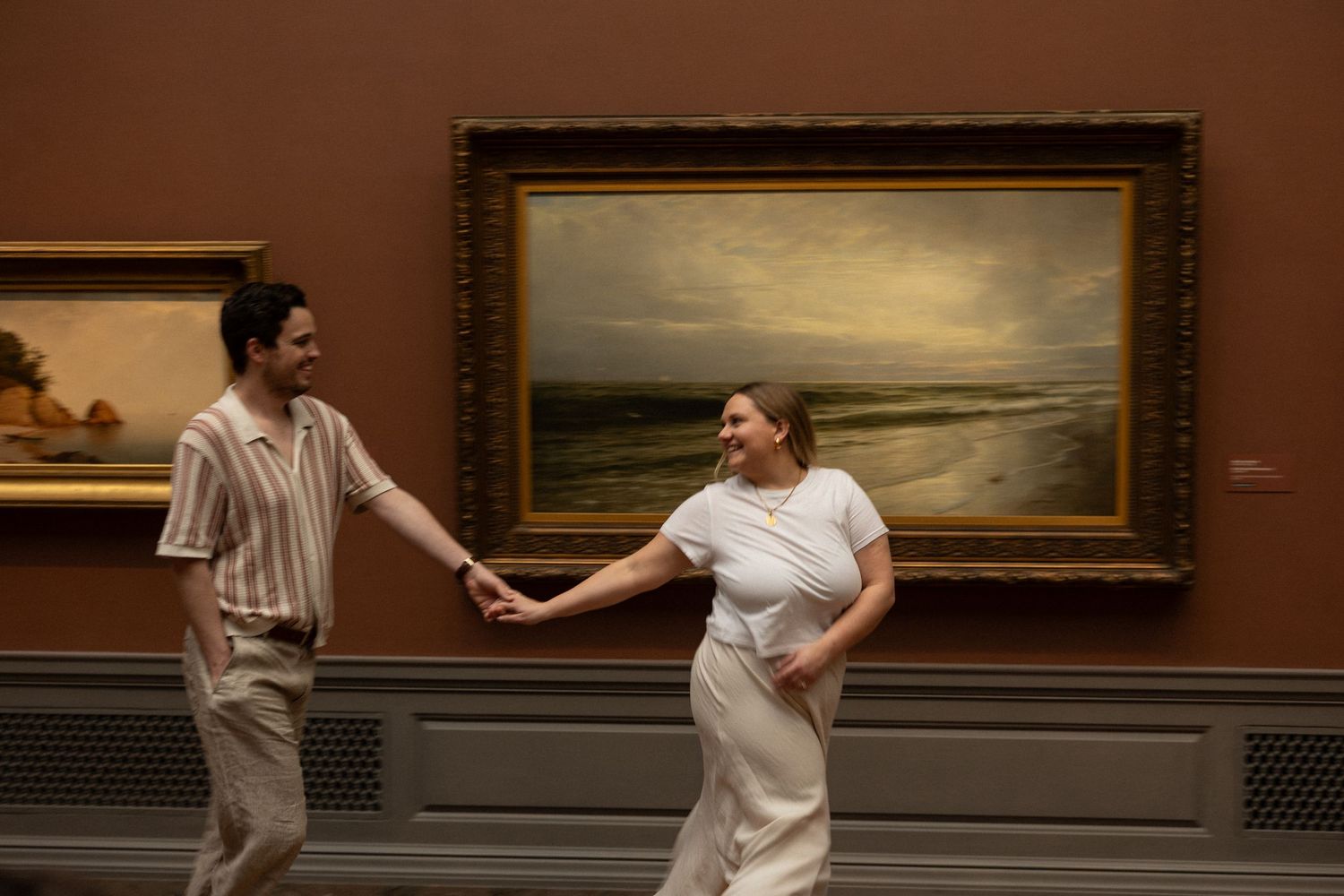 Happy couple holding hands and laughing in front of a large landscape painting in an art museum.