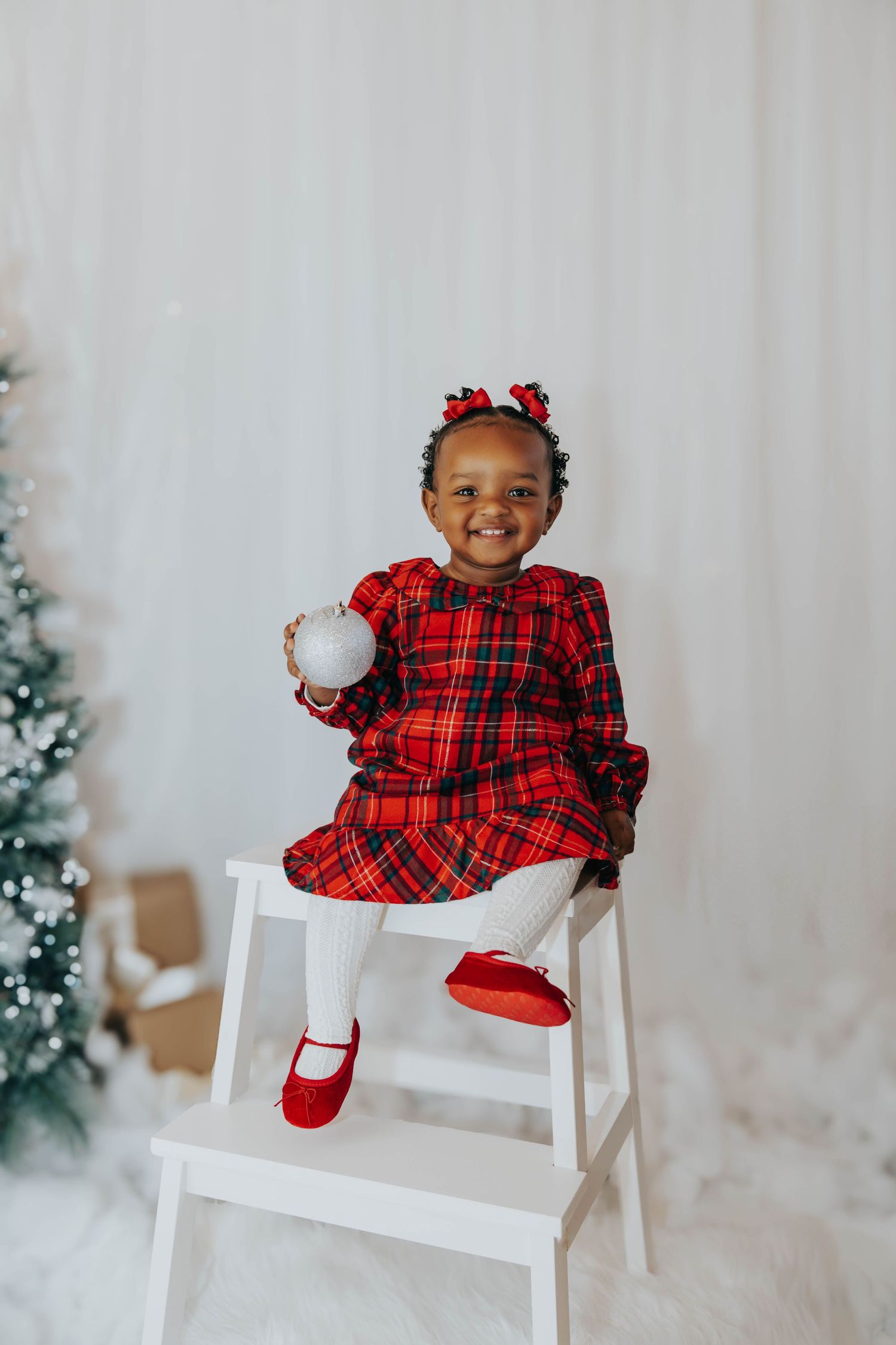Child in plaid red dress and matching hair bows sits on white steps near Christmas tree.