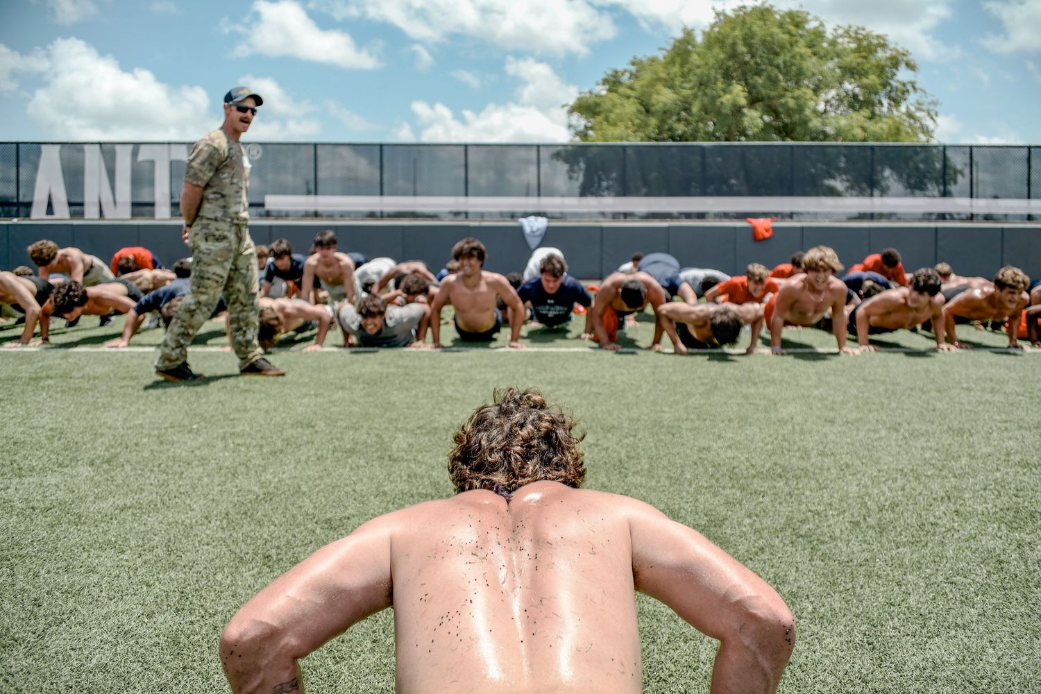 Military training session shows recruits doing pushups on a sports field while an instructor supervises.